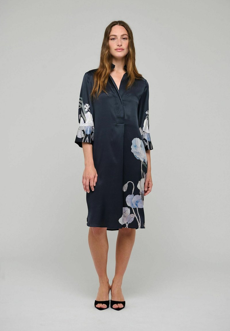 Femme debout portant une robe sombre mi-longue ornée de motifs floraux clairs sur les manches et l'ourlet, associée à des talons noirs ouverts à l'avant.