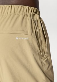 Beige shorts made of lightweight fabric, featuring an elastic waistband, side pockets, and a white "snow peak" logo. Smooth texture.