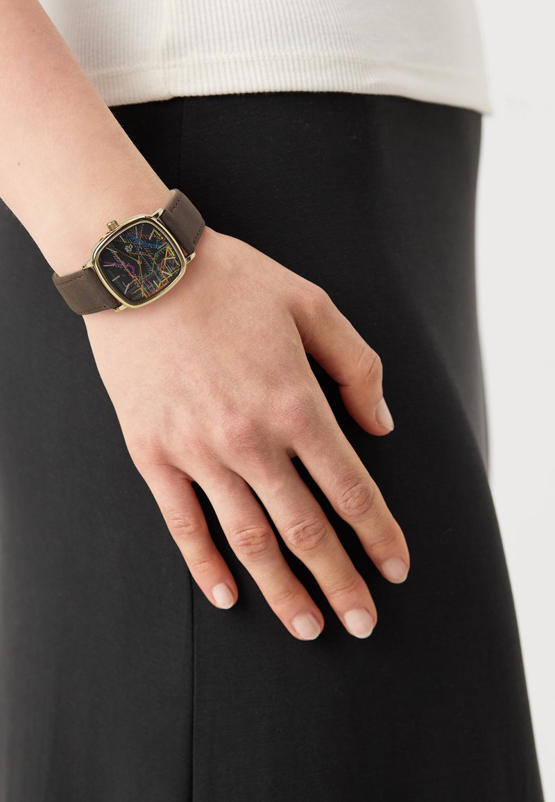 Reloj de pulsera cuadrado con una esfera negra que presenta líneas coloridas y una caja de metal dorada; correa de cuero marrón sujeta en una mano.