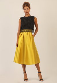 Schwarzes Spitzen-Crop-Top mit gewellten Kanten, kombiniert mit einem fließenden gelben satin Midi-Rock mit Falten und schwarzen High-Heels.