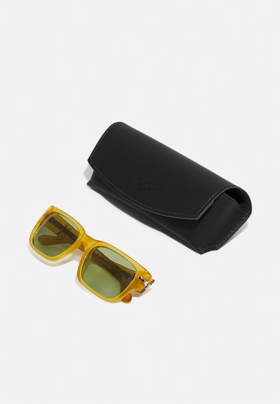 Occhiali da sole gialli a forma rettangolare con lenti verdi accanto a un astuccio in pelle nera. L'astuccio presenta un flap arrotondato e cuciture a contrasto.