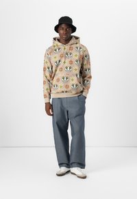 Uomo che indossa un felpa beige con motivo, un cappello a secchiello nero, pantaloni larghi blu e sneakers bianche, in piedi contro uno sfondo bianco semplice.