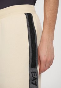 Pantalones jogger crema con una raya negra y etiqueta "EA7", con una textura suave y cinturilla elástica, vistos de lado con una mano.