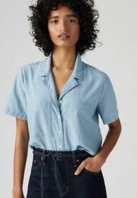 Light blue short-sleeve shirt with a notched collar, two chest pockets, and button front closure, paired with dark jeans and a belt.