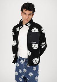 Black fleece jacket with white dice pattern, zippered front. Worn over a white shirt and paired with blue jeans featuring a matching dice print.