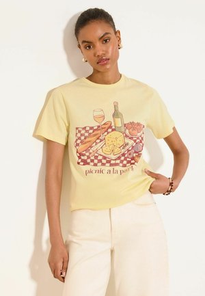 Femeie purtând un tricou galben pal cu o grafică de picnic care arată vin, brânză, baghetă, roșii și textul „picnic a la park.”