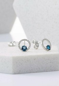 Silberne Steckerohrringe mit runden blauen Steinen, umgeben von kleinen klaren Kristallen, in einem kreisförmigen Design auf einem hellen Stein-Hintergrund.