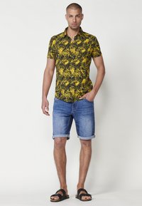 Cămașă cu mâneci scurte și guler de culoare galbenă, cu imprimeu tropical negru, asortată cu pantaloni șort din denim și sandale negre. Pantalonii scurți sunt ruliți la margine.