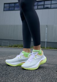 Valkoiset urheilukengät neonvihreillä ja oransseilla yksityiskohdilla, hengittävä verkko-osa, pintakuvioitu pohja, yhdistetty mustiin leggingseihin ja neonvärisiin sukkiin.