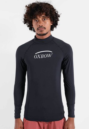 Homme aux cheveux bouclés portant un rash guard noir à manches longues avec le logo "OXBOW" et un short de bain rouge, debout sur fond blanc.