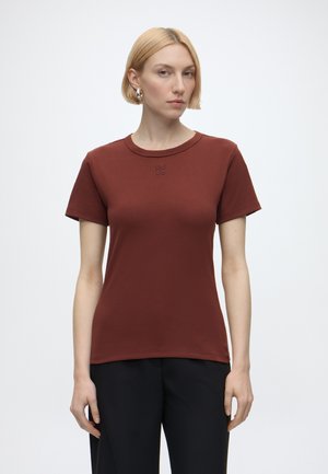 Blond kvinde iført en ensfarvet brun kortærmet t-shirt med teksten "HUGO" på brystet og sorte bukser mod en hvid baggrund.