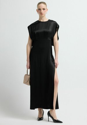 Zwarte satijnen maxi-jurk met korte mouwen en een zijschuif. Gecombineerd met zwarte hoge hakken en een beige handtas. Soepele textuur, elegant ontwerp.
