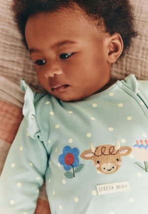 Dojenček leži, oblečen v svetlo zeleno obleko z belimi pikami in vezenimi cvetovi ter nasmejanim obrazom krave z oznako "mali muu".