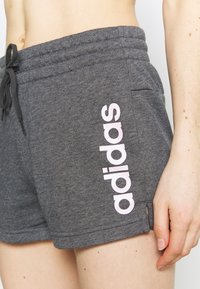 Pantalones cortos deportivos grises con una cinturilla elástica y cordón, con un logo rosa impreso verticalmente en el lado. Textura de tela suave.
