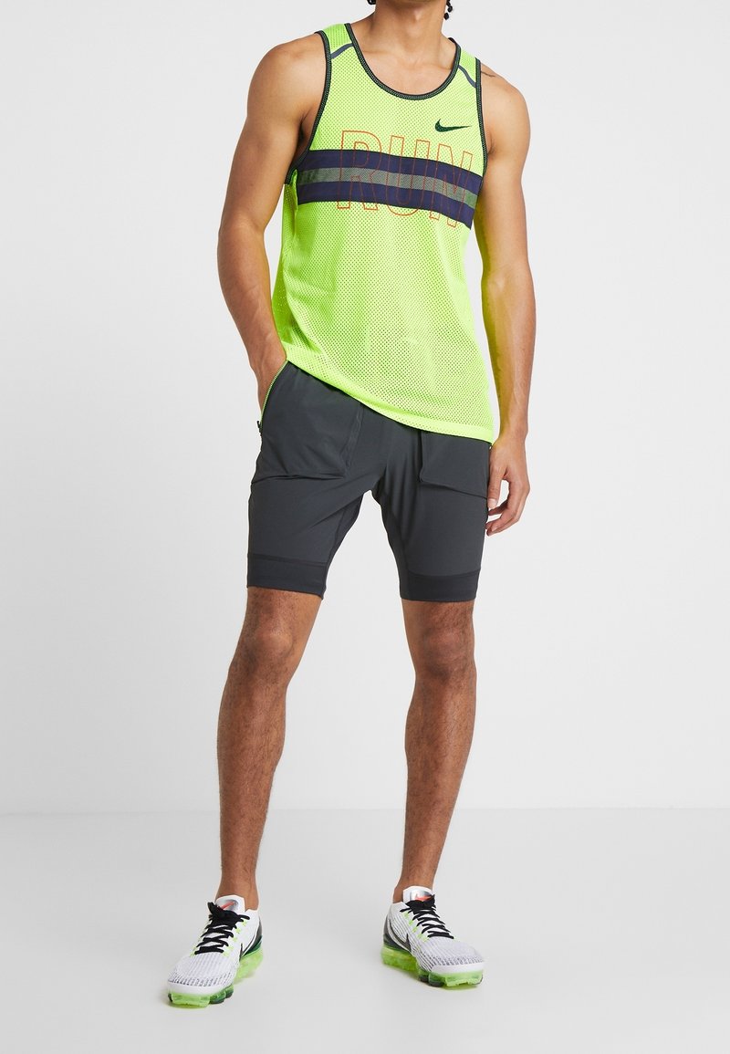 Man som bär en ljusgul ärmlös löpartröja med texten "RUN", mörkgrå shorts med fickor och vita löparskor med gröna sulor.