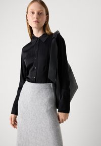 Kvinde iført en sort skjorte med knapper og en grå strikket nederdel, der bærer en stor sort skuldertaske, stående mod en ensfarvet lys baggrund.