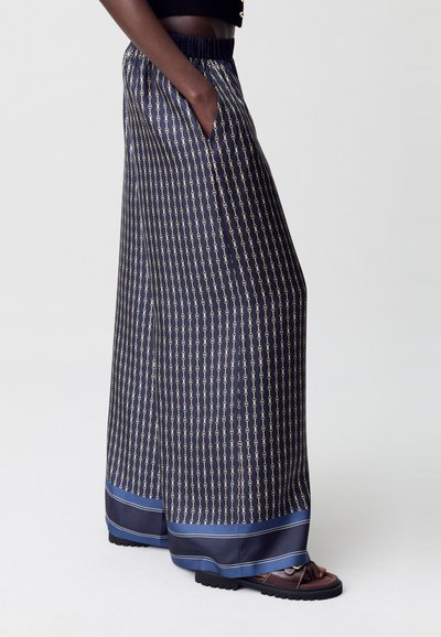 Modelo vistiendo pantalones anchos de patrón en color azul marino con una cintura elástica, con una mano en el bolsillo y combinados con sandalias. Fondo sencillo.
