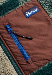 Brown fabric pocket with a blue zipper, featuring a textured green fleece lining and a Penfield logo tag on the edge.