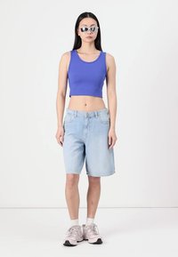 Gekürztes blaues Tanktop kombiniert mit hellblauen Denim-Bermuda-Shorts. Das Model trägt Sonnenbrille mit reflektierenden Gläsern und helle Sneaker mit weißen Socken.