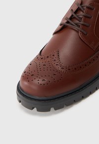 Barna bőr cipő, brogue mintázattal, perforált részletekkel és fekete gumitalppal a tapadásért. A fűző szürke.