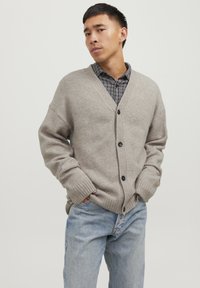 Cardigan beige lavorato a maglia con scollatura a V, chiusura a bottoni e polsini a coste, abbinato a una camicia a scacchi bianca e nera e jeans blu.