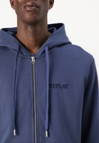 Mørkeblå zip-up hoodie lavet af blødt stof har en hætte med snørebånd, "REPLAY" broderi og sølvdetaljer på lynlåsen.