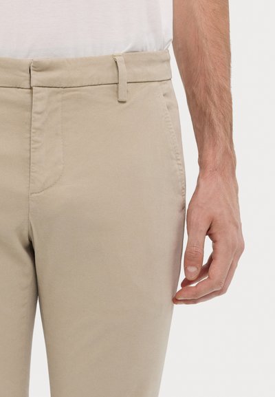 Chino en coton beige avec une texture lisse, coupe ajustée, dotés de passants de ceinture et de poches latérales. Main visible au niveau de la taille.