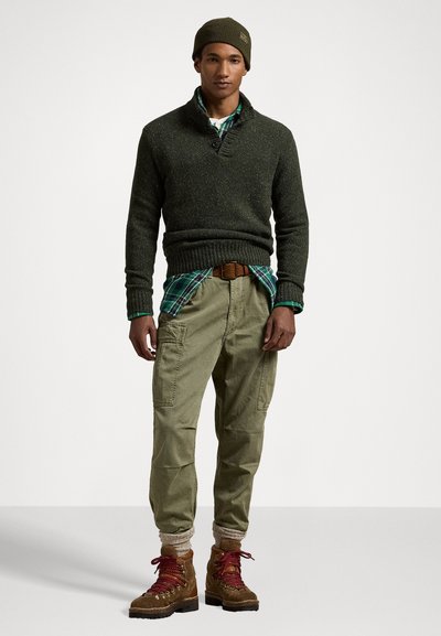 Maglione lavorato a maglia verde scuro con colletto a scialle, indossato sopra una camicia a quadri blu e verde, abbinato a pantaloni cargo color khaki chiaro e stivali marroni con lacci.