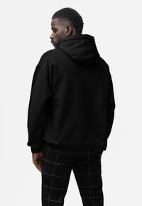 Felpa nera con cappuccio e finitura testurizzata, indossata con pantaloni a quadri bianchi e neri, che mostra una vestibilità comoda e un design casual.