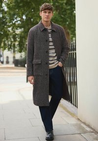 Lang brun struktureret frakke med krave, knaplukning foran og lommer, båret over en mønstret sweater, parret med mørkeblå jeans og sorte sko.