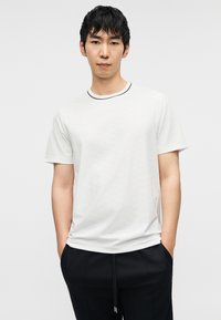 Λευκό κοντομάνικο μπλουζάκι με υφή στην επιφάνεια και μαύρο στρογγυλεμένο ντεκολτέ, φορεμένο με μαύρο παντελόνι, τα χέρια στις τσέπες.