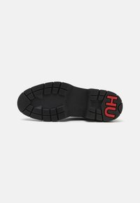 Semelle de chaussure en caoutchouc noir avec un motif de bande de roulement texturé, présentant un logo rouge "HUGO" proéminent sur la zone du talon.