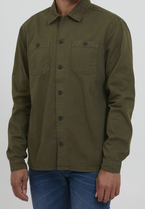 Homme portant une chemise vert olive à boutons avec deux poches poitrine, associée à un jean bleu, debout devant un fond uni.