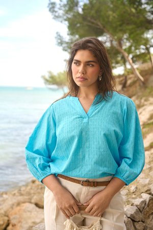Jeune femme en blouse bleue et pantalon beige debout, tenant un sac tissé près d'un rivage rocheux avec des arbres et la mer en arrière-plan.