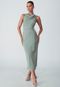 Sleek, mint green sleeveless dress with a cowl neckline, tied at the waist, featuring a midi length and smooth texture. Paired with white strappy heels.