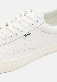 Baskets en cuir blanc au toucher lisse, avec une bande latérale texturée, des lacets blancs et une petite étiquette du logo Vans près de la languette.