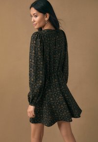 Sort kjole med gyldent blomsterprint, lange pufærmer, indsnøret talje og udvidet kant, lavet af let vævet stof.