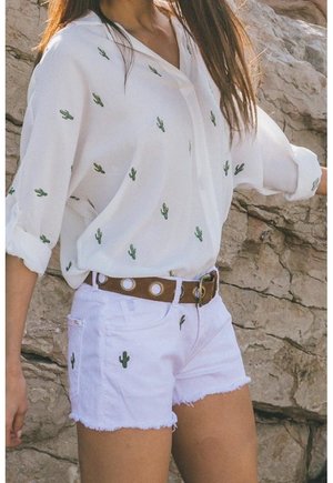 Biała koszula i szorty z małymi zielonymi nadrukami kaktusów, podwinięte rękawy, brązowy pasek z okrągłymi dziurkami, noszone przez osobę stojącą obok skalistej powierzchni.