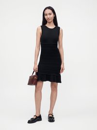 Femme aux longs cheveux foncés portant une robe noire sans manches, des mocassins noirs et tenant un petit sac à main marron sur fond blanc.