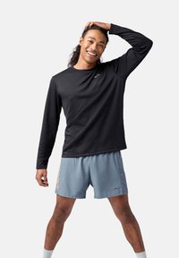 Schwarzes langärmliges Sportshirt mit texturiertem Muster, kombiniert mit hellblauen Shorts, die ein oranges Logo aufweisen. Das Modell präsentiert sich in Pose.