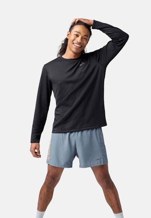 Schwarzes langärmliges Sportshirt mit texturiertem Muster, kombiniert mit hellblauen Shorts, die ein oranges Logo aufweisen. Das Modell präsentiert sich in Pose.