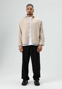 Lichtbeige overhemd met knopen, losse pasvorm, gedragen over een wit T-shirt, gecombineerd met zwarte wijde broekspijpen en zwarte chunky schoenen. Glad oppervlak.