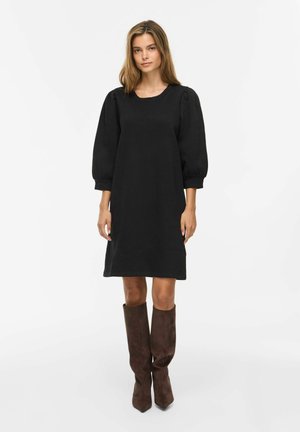 Jeune femme portant une robe noire longueur genou avec des manches trois-quarts bouffantes et des bottes en daim marron montant jusqu'aux genoux, debout sur un fond blanc.