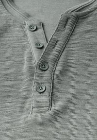 Harmaa teksturoitu henley-paita, jossa on kolme nappia kauluksessa. Kankaassa on raidallinen kuvio, joka lisää sen tuntua.