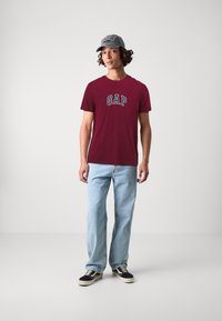 T-shirt GAP bordeaux, jeans bleus, et sneakers noirs. Casquette grise avec texte brodé. Design simple, coupe décontractée, texture de tissu naturel.