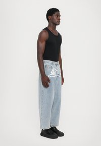 Top preto de alças com jeans de denim azul claro de corte solto, apresentando um design gráfico branco e um aplique em formato de arco-íris na cintura; sapatos pretos.