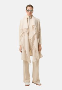 Beżowy szal z frędzlami noszony na dopasowanym golfie i spodniach, zestawiony z białymi butami. Gładka faktura i warstwowy design tworzą spójny wygląd.