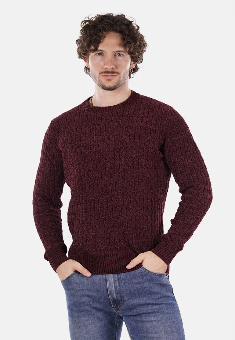 Uomo con capelli ricci che indossa un pullover a treccia borgogna e jeans blu, in piedi con le mani in tasca su uno sfondo bianco.