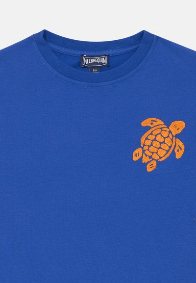Modré tričko s crew-neck, na kterém je na levé části hrudi vyšitá oranžová mořská želva a uvnitř límce je štítek značky Vilebrequin.