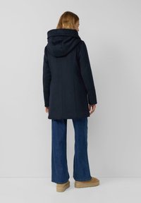 Frau mit langem Haar, die einen marineblauen Kapuzenmantel, blaue Jeans mit weitem Bein und beige Wildleder-Stiefeletten trägt, steht vor einem schlichten weißen Hintergrund.